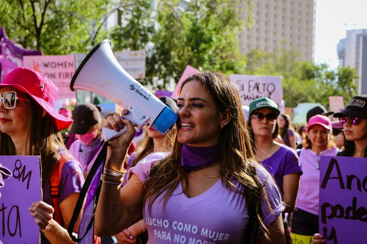 The Art of Drawing Readers In: Your attractive post title goes here A woman passionately leading a protest, speaking into a megaphone with a vibrant crowd in the background.